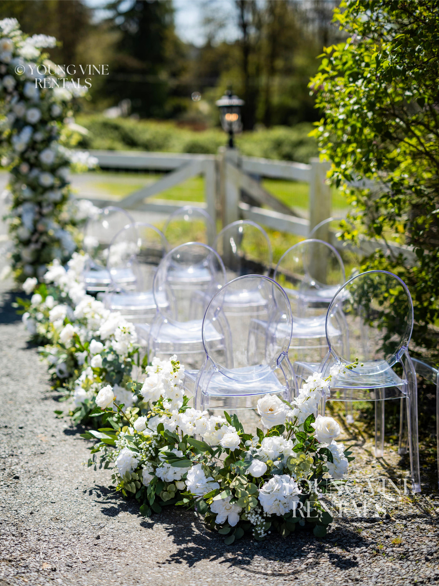 Outdoor winery wedding ceremony with clear acrylic Ghost chairs arranged in four rows. The aisle is decorated with lush floral arrangements in white, cream, and green. In the background is a lush and romantic floral arbor, studded with an abundance of ivory and white flowers and various types of eucalyptus foliage.
