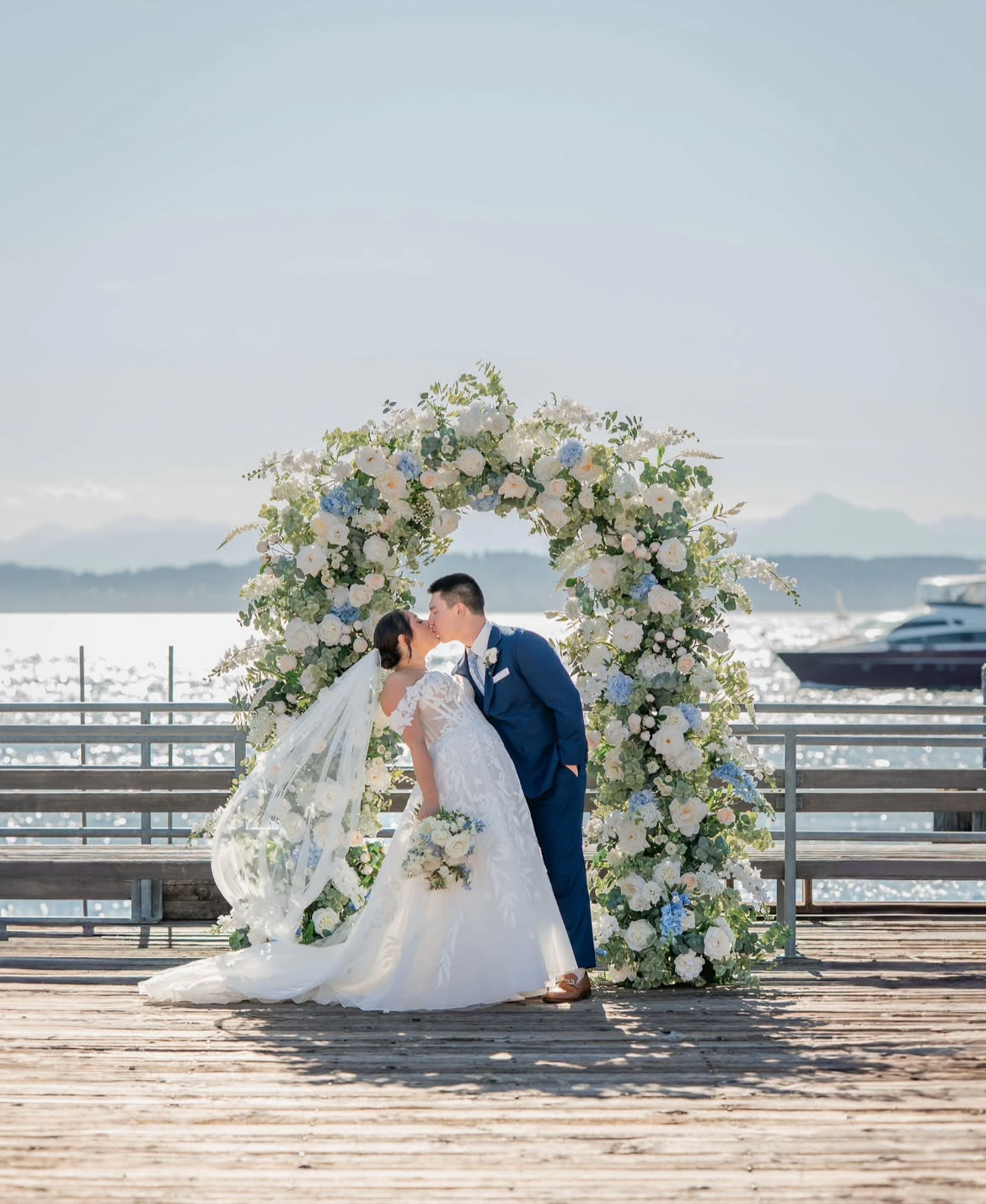 Wedding couple standing under a floral arch on a dock with water in the background.