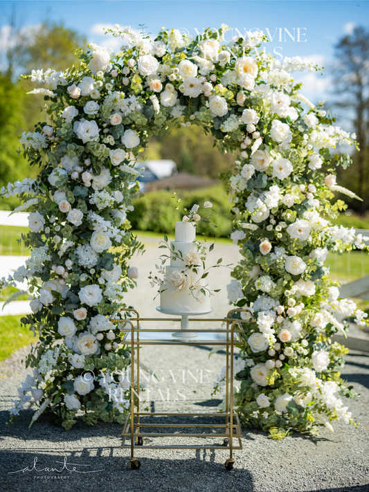 Wedding cake with ivory and green sugar flowers displayed on a gold bar cart, in front of a lush floral arbor at an outdoor winery wedding. The wedding arch in the background is studded with an abundance of ivory and white flowers and various types of eucalyptus foliage.