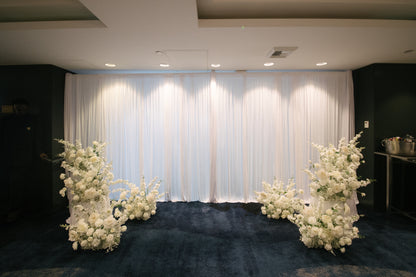 Decorative setup with white floral arrangements on a dark carpeted floor against a white curtain backdrop.